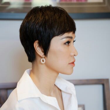 A side profile of a woman wearing a collared shirt and dangling hoop earrings with fine hair and a pixie cut