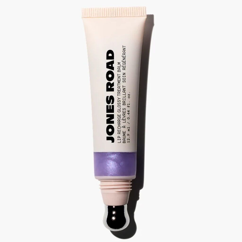 Jones Road Lip Recharge Glossy Treatment Balm in a soft beige tube with a translucent window showing a lilac-tinted formula and a metal applicator tip against a light gray background.