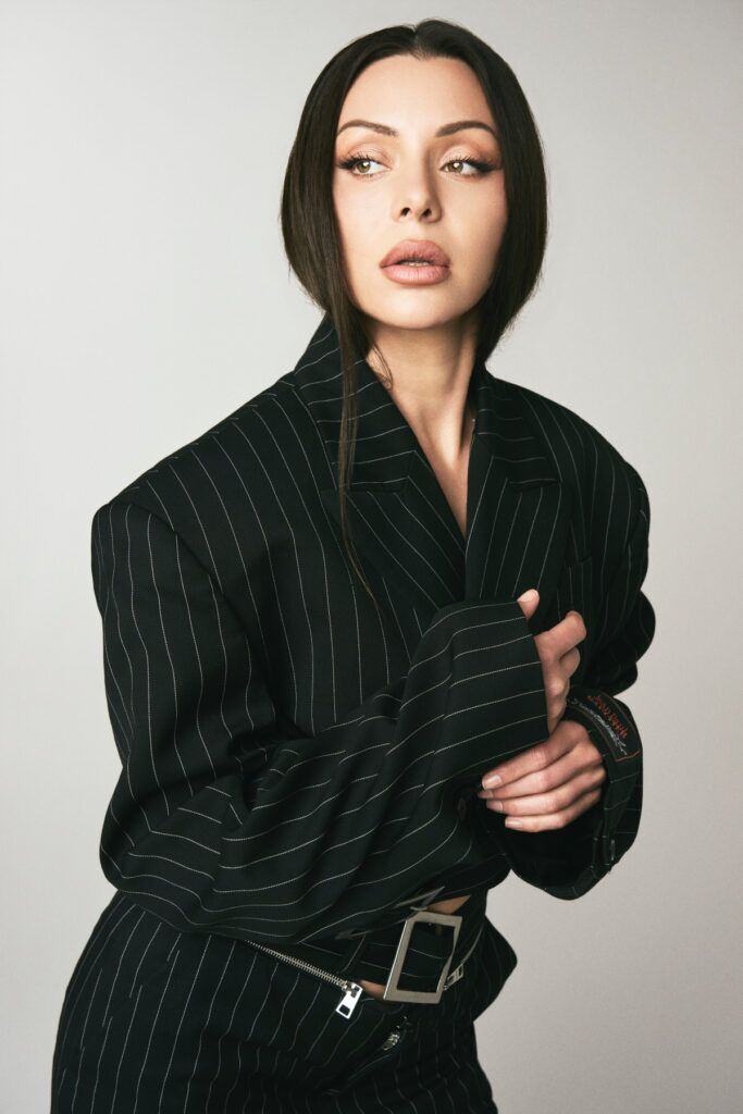 Portrait of Neraida Bega in a black tailored suit, posing in profile with a sleek high ponytail and soft glam makeup