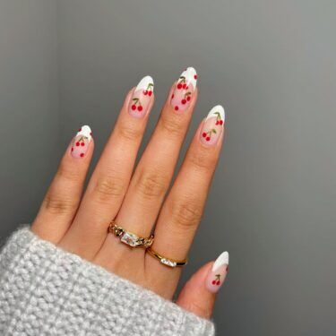 A close up photo of a woman's hand with manicured nails with a cherry design on each nails and two rings on her fingers